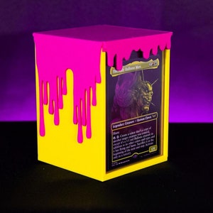 Puede incluir: Una caja amarilla para guardar cartas con un diseño de pintura rosa goteando en la parte superior y lateral. La caja contiene una carta de Magic: The Gathering con el personaje "The Deadly Balloon Man".