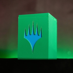 K&ouml;nnte beinhalten: Eine gr&uuml;ne Kartenaufbewahrungsbox mit einem blauen Emblem. Die Box hat eine W&uuml;rfelform und eine strukturierte Oberfl&auml;che. Das Emblem ist ein stilisiertes Design mit kronenartiger Form. Die Box ist f&uuml;r Sammelkartenspiele konzipiert.