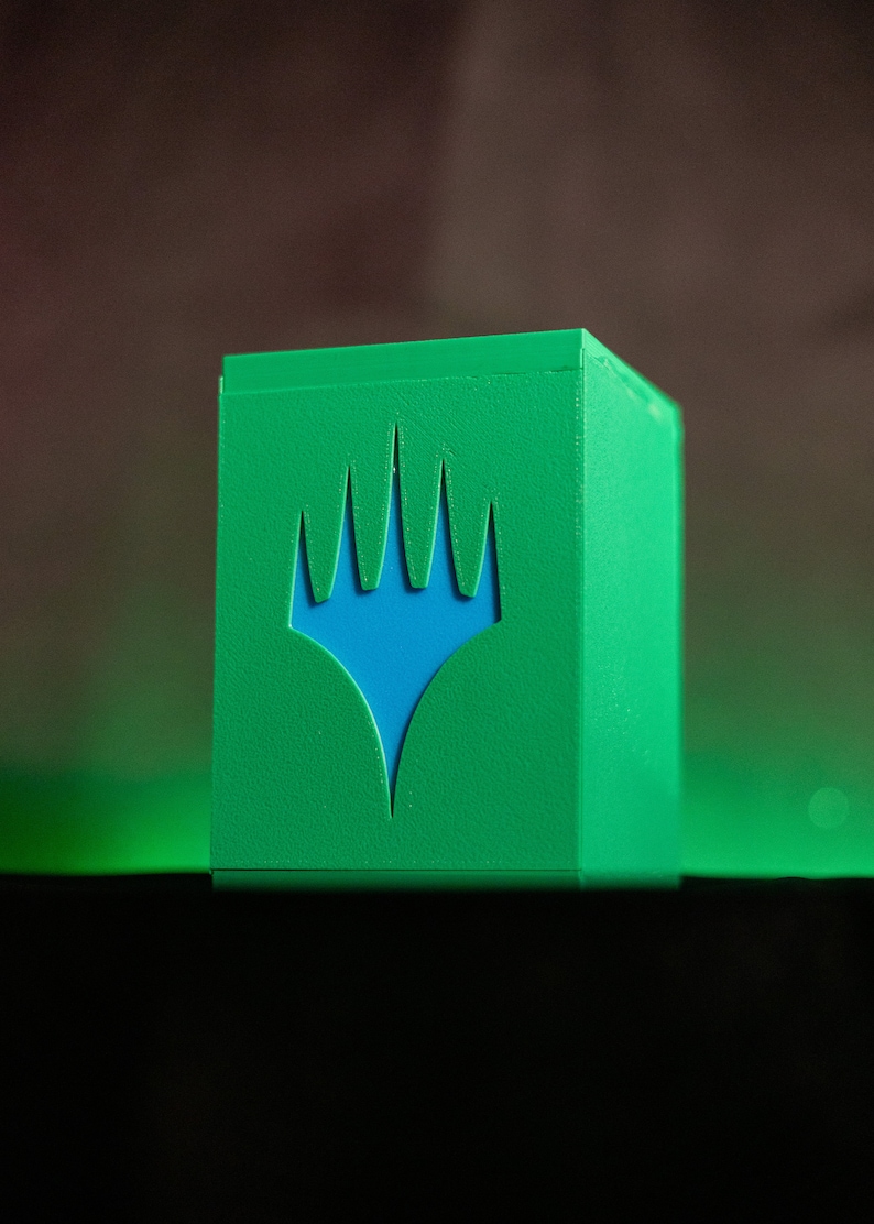 K&ouml;nnte beinhalten: Eine gr&uuml;ne Kartenaufbewahrungsbox mit einem blauen Emblem. Die Box hat eine W&uuml;rfelform und eine strukturierte Oberfl&auml;che. Das Emblem ist ein stilisiertes Design mit kronenartiger Form. Die Box ist f&uuml;r Sammelkartenspiele konzipiert.