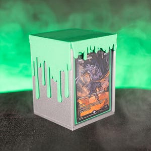 Könnte beinhalten: Kartenaufbewahrungsbox mit grünem, tropfendem Schleimdesign auf silberner Basis. Die Box enthält eine Sammelkarte mit einer Fantasy-Illustration.