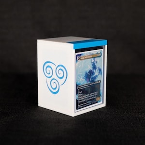 Op de afbeelding: Een witte kaartenopbergdoos met een blauwe rand en een blauw lucht-nomadensymbool. De doos bevat een kaart met de tekst "Aang, Swift Savior". De kaart heeft een blauwe karakterillustratie.