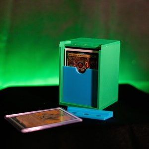 K&ouml;nnte beinhalten: Eine gr&uuml;ne und blaue Kartenaufbewahrungsbox mit einer Karte mit der Aufschrift "Volo, Guide to Monsters". Eine Karte in einer Schutzh&uuml;lle und eine blaue Basis befinden sich vor der Box. Der Hintergrund ist ein Farbverlauf von Gr&uuml;n und Schwarz.