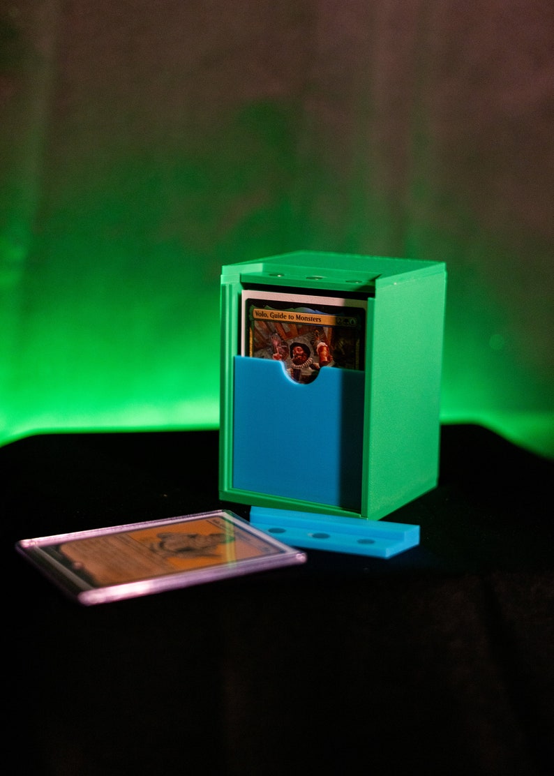 K&ouml;nnte beinhalten: Eine gr&uuml;ne und blaue Kartenaufbewahrungsbox mit einer Karte mit der Aufschrift "Volo, Guide to Monsters". Eine Karte in einer Schutzh&uuml;lle und eine blaue Basis befinden sich vor der Box. Der Hintergrund ist ein Farbverlauf von Gr&uuml;n und Schwarz.