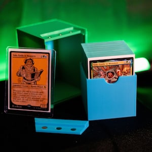 K&ouml;nnte beinhalten: Eine blaue Kartenaufbewahrungsbox mit einem Stapel Karten und einer einzelnen Karte in einer Schutzh&uuml;lle. Auf der Karte steht "Volo, Guide to Monsters". Ein passender blauer Kartenhalter und eine gr&uuml;ne Box sind ebenfalls vorhanden.