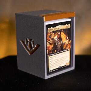 May include: A black and gray card storage box with a gold accent on the top edge. The box has a gold emblem on the side and holds a deck of cards, one of which is visible, featuring the text "Vihaan, Goldwaker."