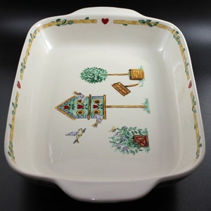 May include: White ceramic baking dish with a green, blue, and red birdhouse design. The dish has a scalloped edge and two handles.