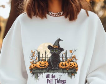May include: White sweatshirt with a black cat wearing a witch hat and holding a pumpkin. The cat is sitting on a fence with pumpkins and the text "All the Fall Things" is below the cat.