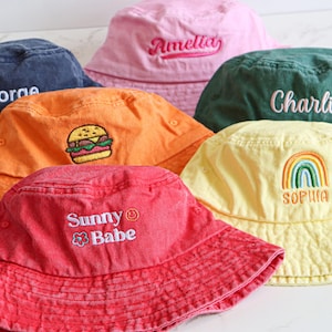 Op de afbeelding: Een verzameling kleurrijke vissershoeden. De hoeden zijn er in verschillende kleuren, waaronder marineblauw, roze, oranje, groen, rood en geel. Elke hoed heeft unieke borduursels, met namen als "George", "Amelia", "Charlie" en "Sophia", en de tekst "Sunny Babe".
