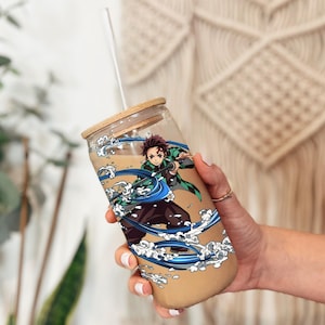 K&ouml;nnte beinhalten: Ein durchsichtiger Glasbecher mit Bambusdeckel und Strohhalm. Der Becher hat ein farbenfrohes Anime-Charakter-Design mit blau-wei&szlig;en Wellenmustern. Der Becher ist mit einer hellbraunen Fl&uuml;ssigkeit gef&uuml;llt.