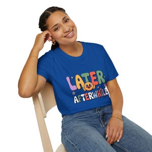 May include: A royal blue t-shirt with the words "LATER AFTERWHILE" in colorful, playful lettering. The design includes a cartoon character and decorative elements. The shirt is made of a soft material.