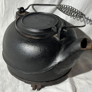 Op de afbeelding: Een zwarte gietijzeren ketel met een metalen handvat en een spiraalvormige grip. De ketel heeft een ronde body, een tuit en een deksel. De ketel staat op een voet met drie poten.