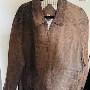 May include: A brown leather jacket with a textured finish, featuring a collared neckline, a front zipper, and two front pockets. The jacket has a checkered pattern lining visible at the collar and a ribbed hem.