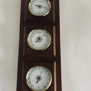 May include: A wooden weather station with three circular gauges. The top gauge reads "Normal", the middle reads "Rain", and the bottom gauge displays temperature in Fahrenheit. A decorative eagle tops the dark brown wooden frame.