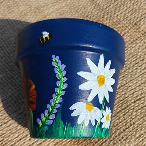 Può includere: Un vaso per piante blu navy con disegni floreali dipinti a mano che includono margherite bianche con centri gialli, steli di lavanda viola e un fiore rosso. Una piccola ape gialla e nera è dipinta vicino al bordo superiore del vaso.