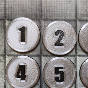 Op de afbeelding: Negen ronde tokens van zilverkleur met zwarte nummers van 1 tot en met 9.
