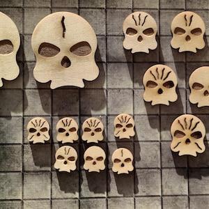 Könnte beinhalten: Eine Sammlung von Holz-Spielsteinen in Schädelform in verschiedenen Größen. Die Schädel sind alle aus hellem braunem Holz geschnitzt und haben schwarze Details. Die Spielsteine sind in einem Gittermuster angeordnet.