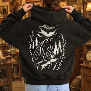 May include: A black hooded sweatshirt featuring a white graphic print on the back. The design depicts a moth-like creature standing on a cliff edge in front of a forest and a large moon, with a smaller flying moth above, all accented by red eyes.