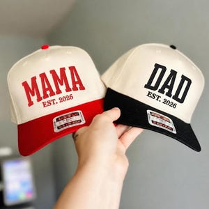 May include: Two baseball caps, one red and cream with "MAMA EST. 2026" in red lettering, and another black and cream with "DAD EST. 2026" in black lettering. Both caps have a structured design and a flat brim.