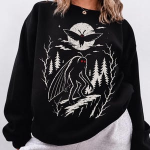 May include: A black long-sleeve sweatshirt featuring a white graphic print of a winged creature with glowing red eyes standing on a cliff. The design includes a full moon, pine trees, and bare branches, creating a dark, forest-themed illustration.