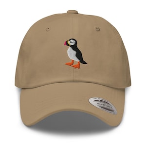 May include: A tan baseball cap featuring an embroidered puffin bird design. The puffin has a black and white body, orange feet, and a colorful beak. The cap has an adjustable strap and a curved brim.