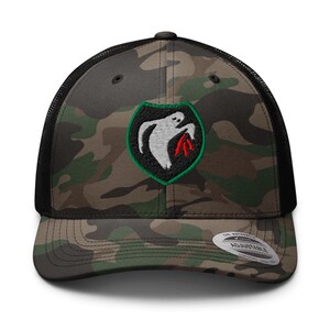 May include: Camouflage trucker hat with a black mesh back and a curved brim. The front features a patch with a white ghost design holding red flames within a green and black shield. The hat is adjustable.