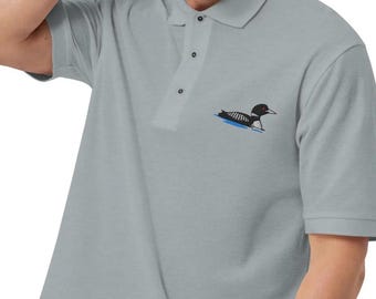 May include: A light gray polo shirt with short sleeves and a collar. The shirt features a small embroidered design of a black and white loon swimming on blue water. The shirt has three buttons and a classic polo design.