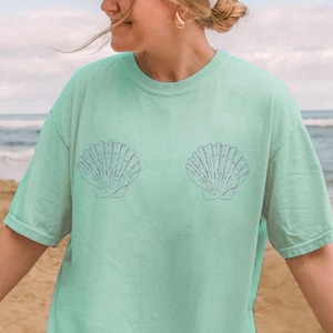 May include: A light mint green short-sleeve t-shirt featuring two dark green line-art seashell illustrations on the chest. The shirt is paired with a plain light beige baseball cap. The text Island Reef is visible in a small circle in the corner.