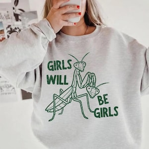 May include: Ash gray sweatshirt with a green graphic of two praying mantises and the text "GIRLS WILL BE GIRLS". The sweatshirt has long sleeves and a crew neck. The word "ASH" is printed in the bottom right corner.