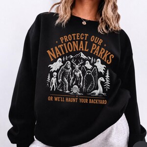 May include: A black crewneck sweatshirt featuring a graphic print in white and orange. The text reads PROTECT OUR NATIONAL PARKS OR WE'LL HAUNT YOUR BACKYARD. The illustration depicts various cryptids, ghosts, and forest scenery in white.