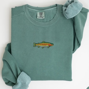 May include: Light green sweatshirt with a crew neck and long sleeves. The sweatshirt features an embroidered fish design in shades of green, orange, and yellow. The cuffs are folded up, revealing a lighter green fabric. The tag reads "Comfort Colors".