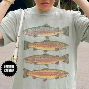 May include: A light sage green short-sleeve t-shirt featuring a graphic print of four stacked rainbow trout fish. The fish are illustrated in shades of green, yellow, and pink with spotted patterns. A black badge with the text ORIGINAL CREATOR is visible.