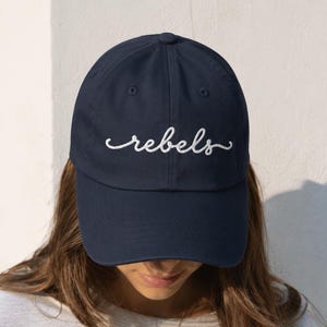 May include: A navy blue baseball cap featuring the word rebels embroidered in white cursive script across the front panel. The cap has a structured crown, a curved brim, and a button at the top center, providing a classic look with clean lines.