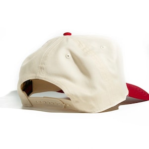 Op de afbeelding: Cr&egrave;mekleurige baseballpet met een rode klep en knoop. De pet heeft een gebogen klep en een verstelbare snapbacksluiting. De pet is gemaakt van een duurzame stof.