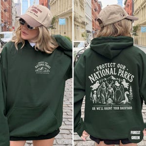 May include: A forest green hooded sweatshirt featuring a small white graphic on the front chest that reads Protect Our National Parks. The back displays a large white graphic of cryptids and ghosts with the text Protect Our National Parks Or We'll Haunt Your Backyard.
