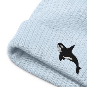 May include: A light blue ribbed knit beanie featuring an embroidered black and white orca whale design on the folded cuff. The hat is constructed with a vertical textured knit pattern and a thick, folded brim that displays the marine animal graphic.