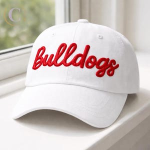 May include: A white baseball cap featuring the word "Bulldogs" embroidered in red script. The cap has a curved peak and a button on the crown.