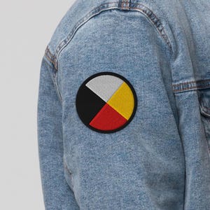 May include: A light blue acid-wash denim jacket sleeve featuring a circular embroidered patch. The patch is divided into four equal quadrants colored white, yellow, red, and black, all enclosed within a thick black circular border.