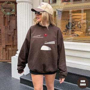 May include: A dark brown sweatshirt with a graphic design featuring white and red accents. The design includes a white shape with vertical lines and a red dot. The sweatshirt is paired with a beige cap and sunglasses.
