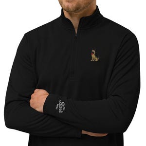 May include: A black quarter-zip pullover featuring a small embroidered German Shepherd dog on the left chest. The right cuff displays the white text "THE BARK NINE" in a cursive script font. The garment has a high collar and long sleeves.