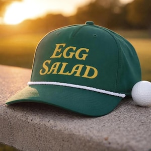 May include: A green baseball cap with the words "EGG SALAD" embroidered in yellow. The cap has a white rope detail along the brim and a white golf ball is next to it.