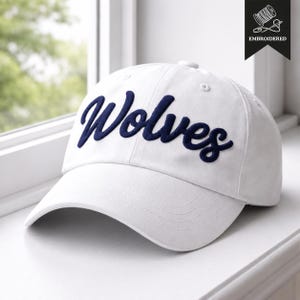 May include: A white baseball cap with the word "Wolves" embroidered in navy blue script across the front. The cap has a curved brim and a button on top. The word "Embroidered" is printed on a black tag.