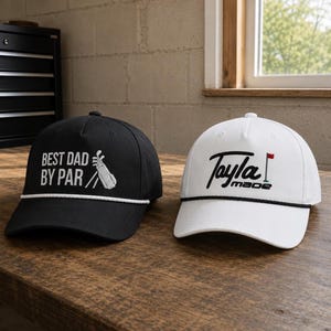 K&ouml;nnte beinhalten: Eine schwarze Baseballkappe mit wei&szlig;em Text BEST DAD BY PAR und einer Golfbag-Grafik sowie eine wei&szlig;e Baseballkappe mit schwarzem Text Tayla made und einer Golffahnen-Grafik. Beide Kappen haben eine dekorative Kordel entlang des Schirms.