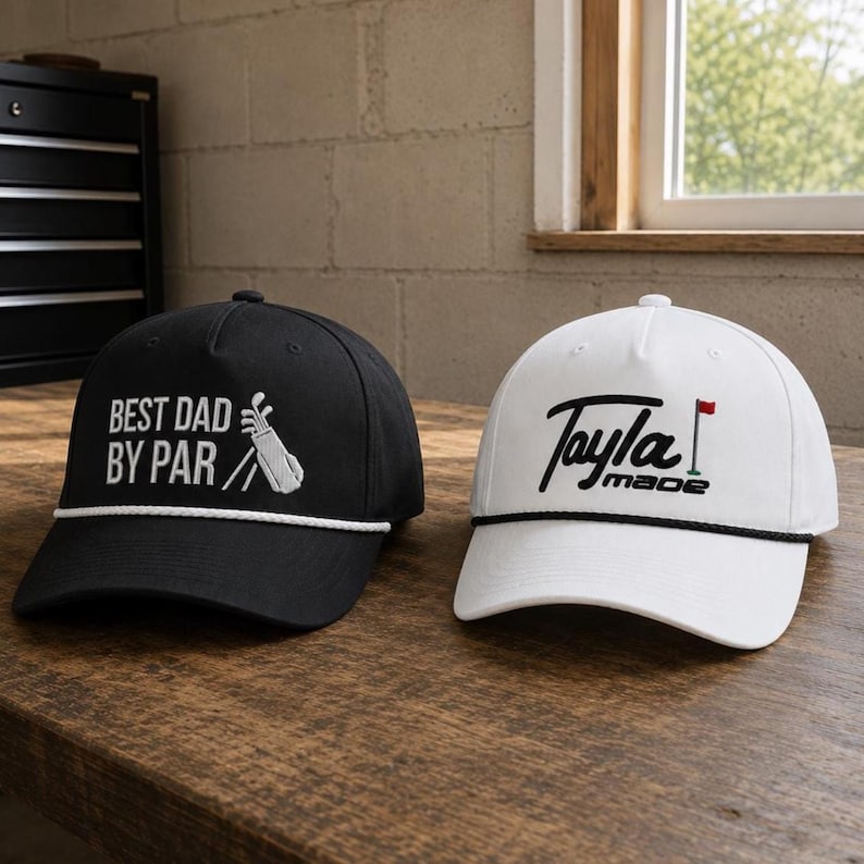 K&ouml;nnte beinhalten: Eine schwarze Baseballkappe mit wei&szlig;em Text BEST DAD BY PAR und einer Golfbag-Grafik sowie eine wei&szlig;e Baseballkappe mit schwarzem Text Tayla made und einer Golffahnen-Grafik. Beide Kappen haben eine dekorative Kordel entlang des Schirms.