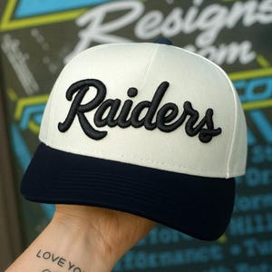 May include: A baseball cap featuring a white crown and a dark navy blue brim. The word Raiders is embroidered in black, cursive, raised lettering across the front panels of the cap. The design includes a structured crown and a curved visor.