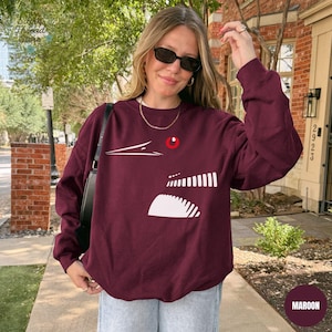 May include: Maroon sweatshirt with a white graphic design. The design features a stylized face with a red circle and white lines. The sweatshirt is a solid maroon color and has a crew neck.