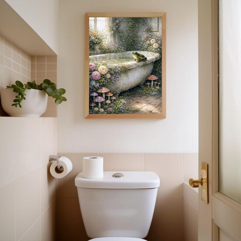May include: A framed art print depicts a whimsical scene of a vintage bathtub surrounded by flowers, mushrooms, and a frog. The artwork is in a bathroom setting, above a white toilet and next to a shelf with a potted plant. The frame is a light brown color.