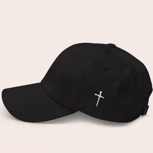 May include: A black baseball cap with a curved brim. A white embroidered cross and nail design is on the side of the cap. The cap has an adjustable strap at the back.