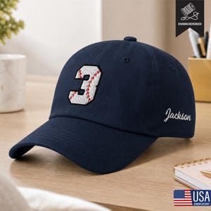 May include: A navy blue baseball cap featuring an embroidered white number 3 with red baseball-style stitching details on the front. The side of the cap displays the name Jackson in white cursive script embroidery.