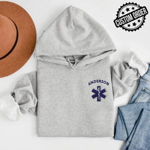 May include: A light grey hooded sweatshirt featuring a dark blue embroidered Star of Life medical symbol on the left chest. The name ANDERSON is stitched in dark blue capital letters above the symbol. The hoodie has long sleeves and a hood.