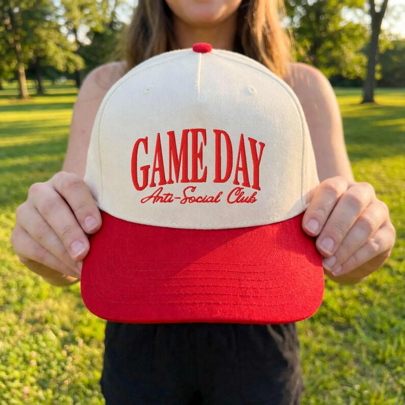 Op de afbeelding: Een baseballpet met een rode klep en een cr&egrave;mekleurige kroon. De pet heeft de woorden "GAME DAY" en "Anti-Social Club" in rode geborduurde letters. De pet heeft een rode knop bovenop.